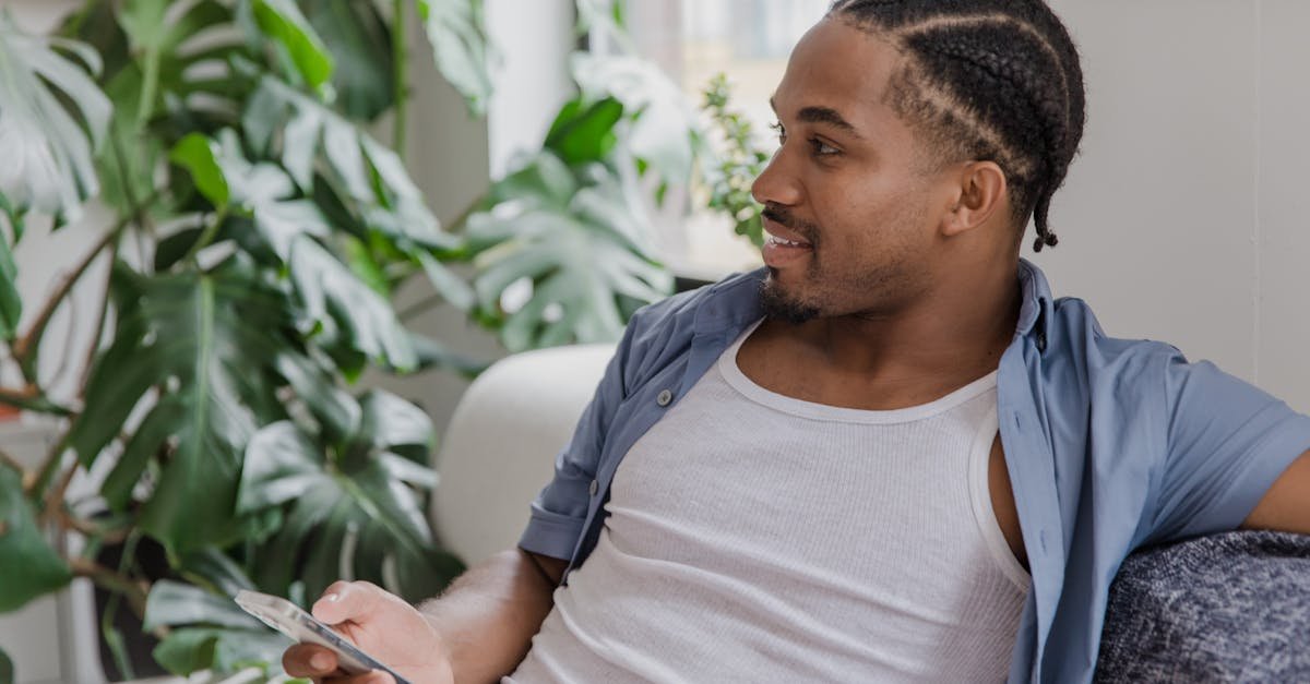 A smiling man with braided hair using his smartphone while sitting on a sofa at home.