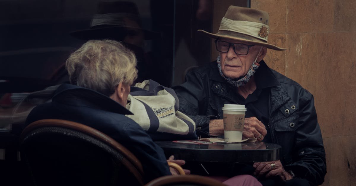 Elderly couple at an outdoor cafe sharing a coffee, wearing hats and jackets.