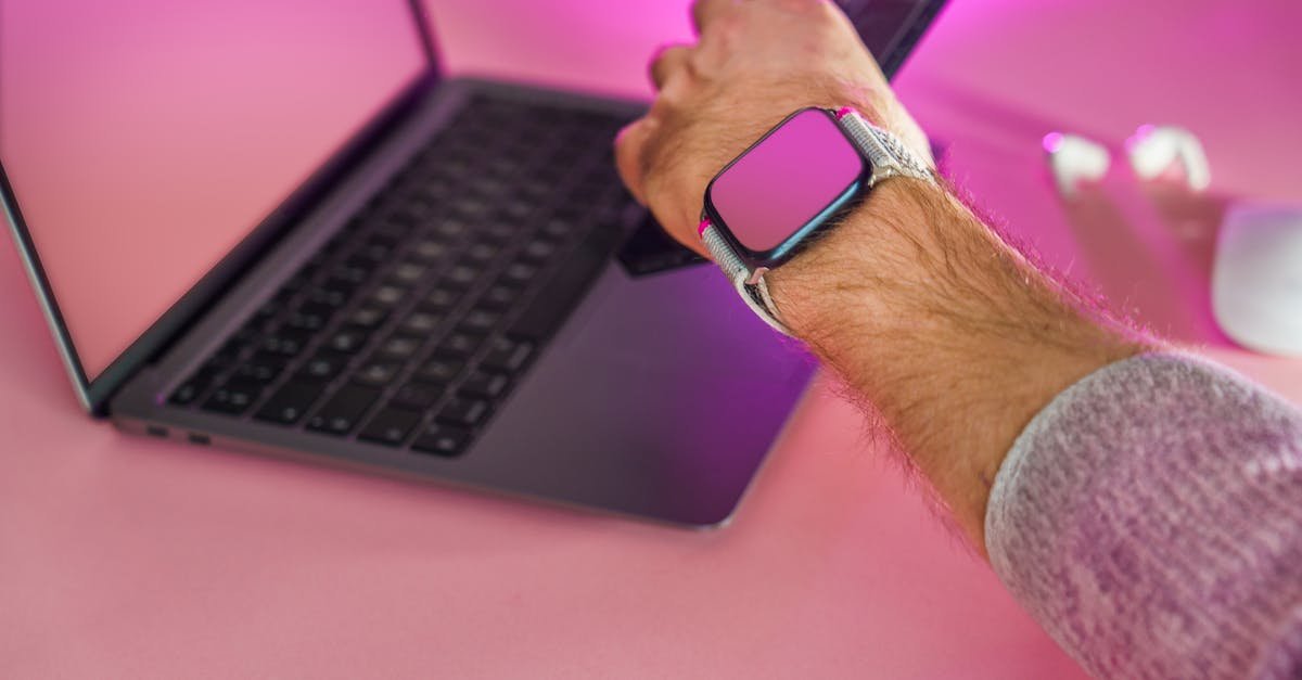Close-up of a hand using a smartphone, smartwatch, and laptop on a pink desk.