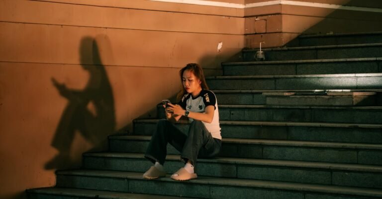 A young woman sits on steps, engaged with her phone. Warm lighting casts a distinct shadow, creating a moody urban scene.