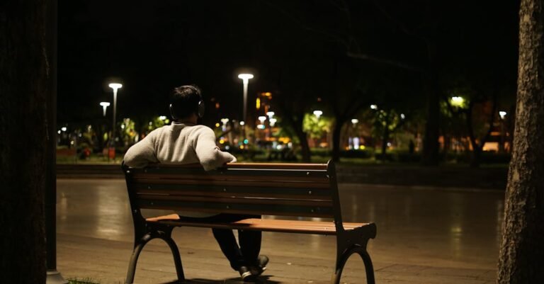 A man with headphones sitting alone on a park bench under lampposts at night.