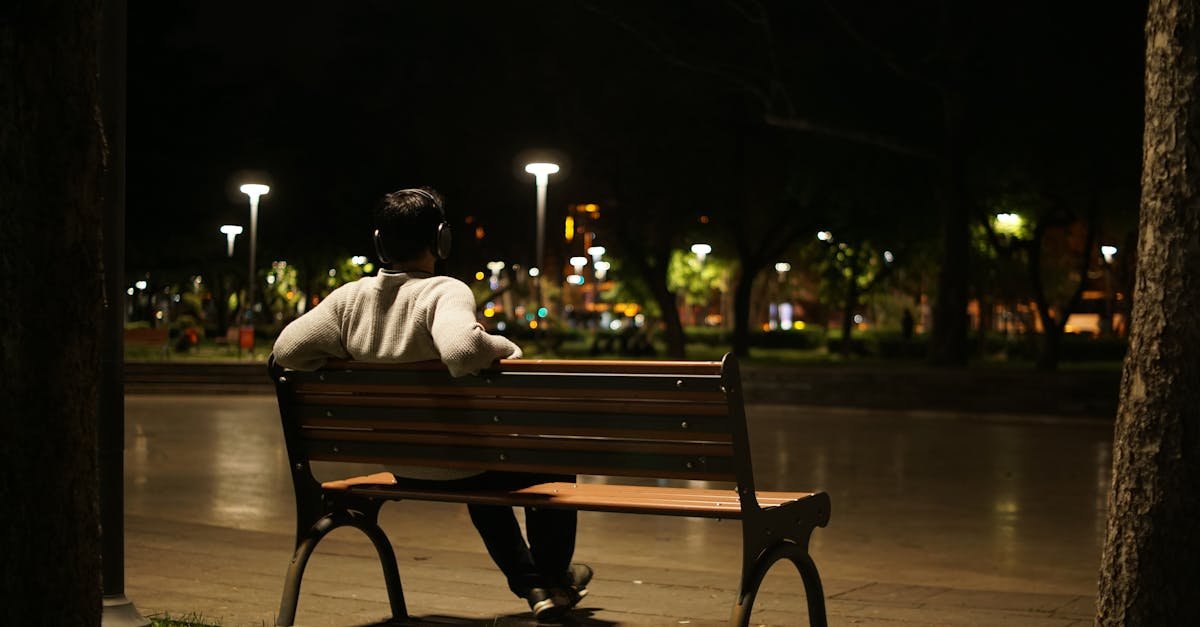 A man with headphones sitting alone on a park bench under lampposts at night.