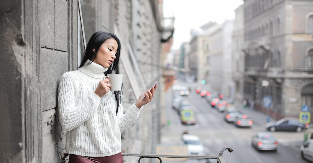 Young woman sipping coffee and using phone on a city balcony.