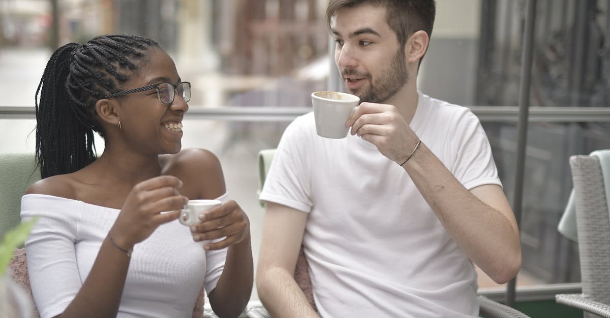 Positive young African American woman in eyeglasses sitting near happy boyfriend while drinking coffee together and looking at each other in cafe