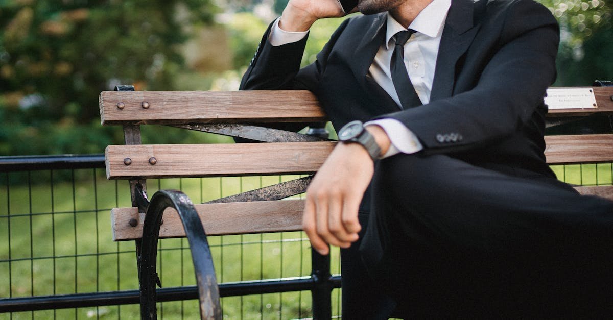 Well-dressed man in a black suit talking on a phone while seated on a park bench during the day.