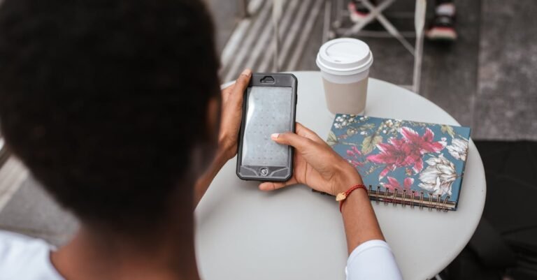 High angle back view of faceless African American lady sitting at table in cafeteria and checking notification on smartphone near notebook and cup of drink