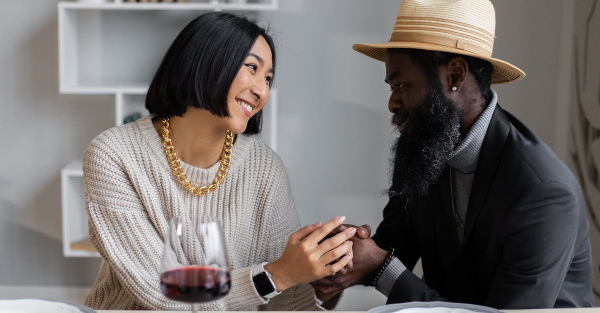 Loving multiracial couple looking at each other holding hands while sitting at table with plates and wine and having dinner