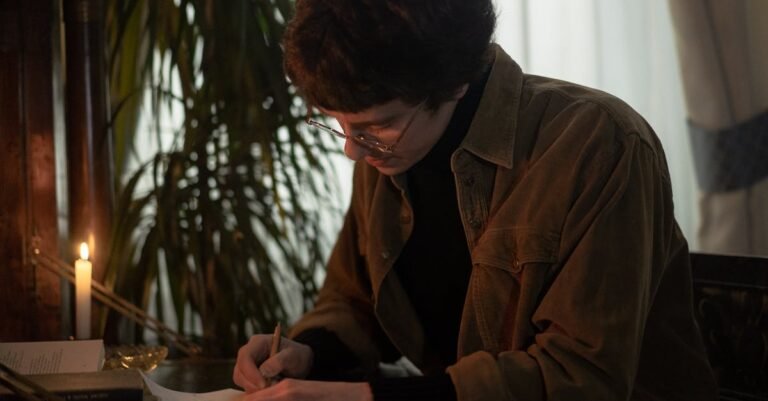 A person in glasses writing at a desk by candlelight, creating a warm ambiance.