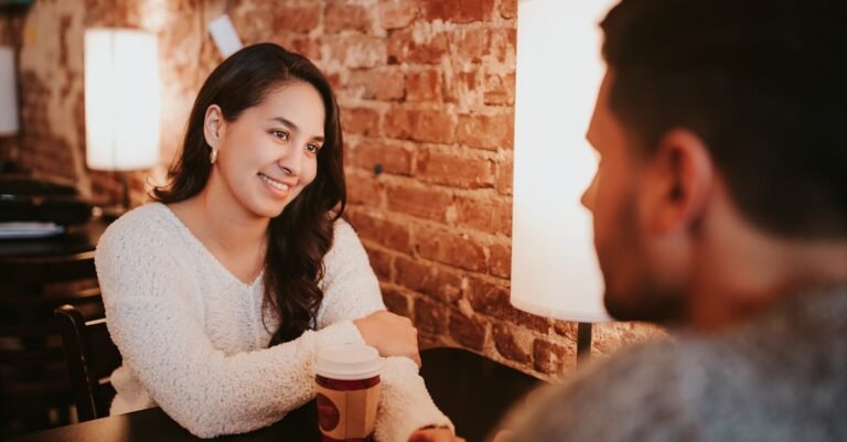 Young couple in casual outfit sitting at table in cafe with cups of drink on date near brick wall and holding hands