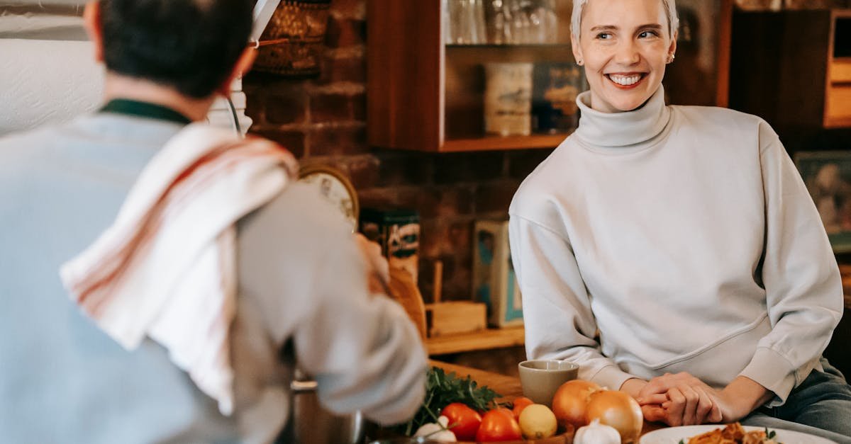 Smiling adult couple spending time in light kitchen while male cooking meal on counter with plates with pasta near onions and tomatoes and herbs near female