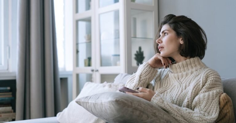 Woman in a comfy sweater sitting on couch, looking thoughtful with a smartphone in hand.