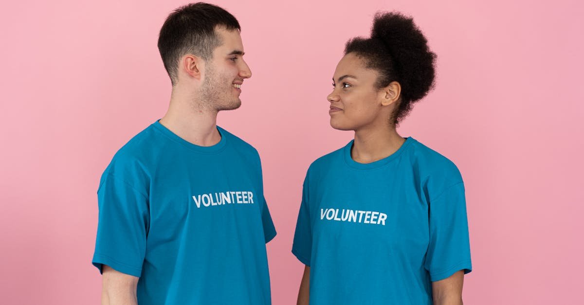 Two diverse volunteers smiling at each other, wearing blue shirts with 'VOLUNTEER' text against a pink background.