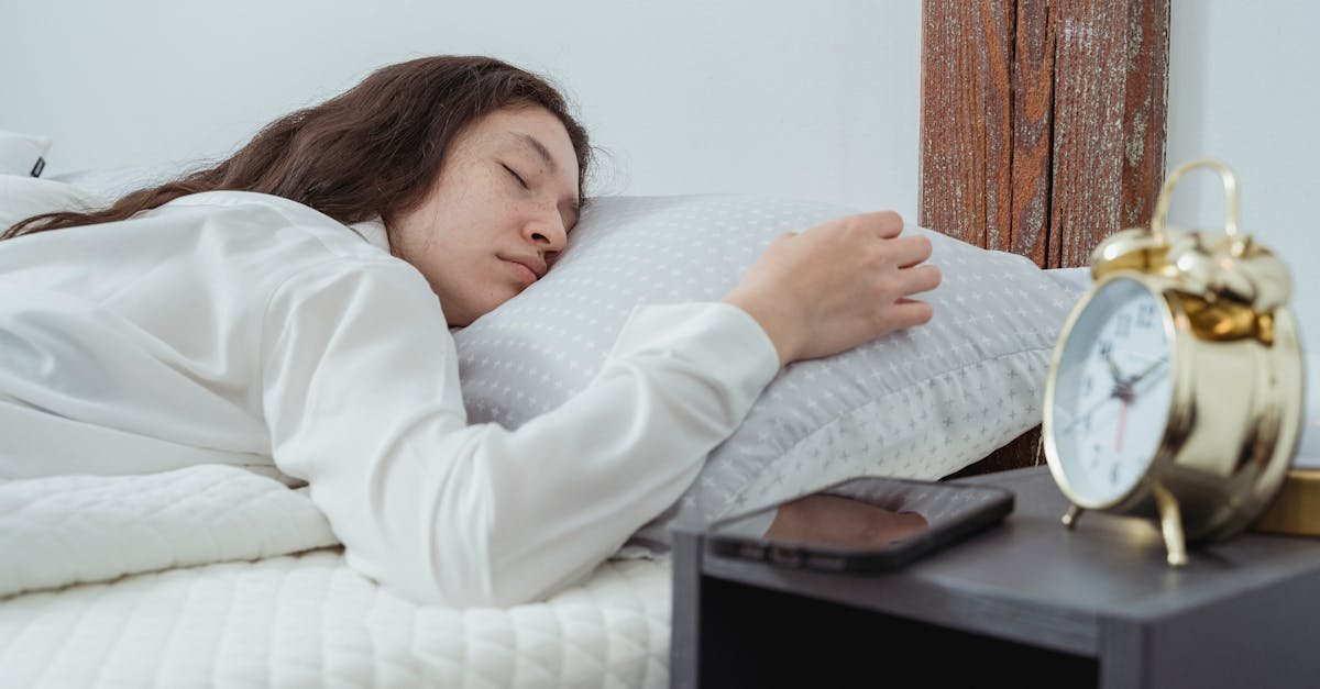 Young woman with dark long wavy hair sleeping peacefully on belly on comfortable bed under white blanket near bedside table with alarm clock and smartphone