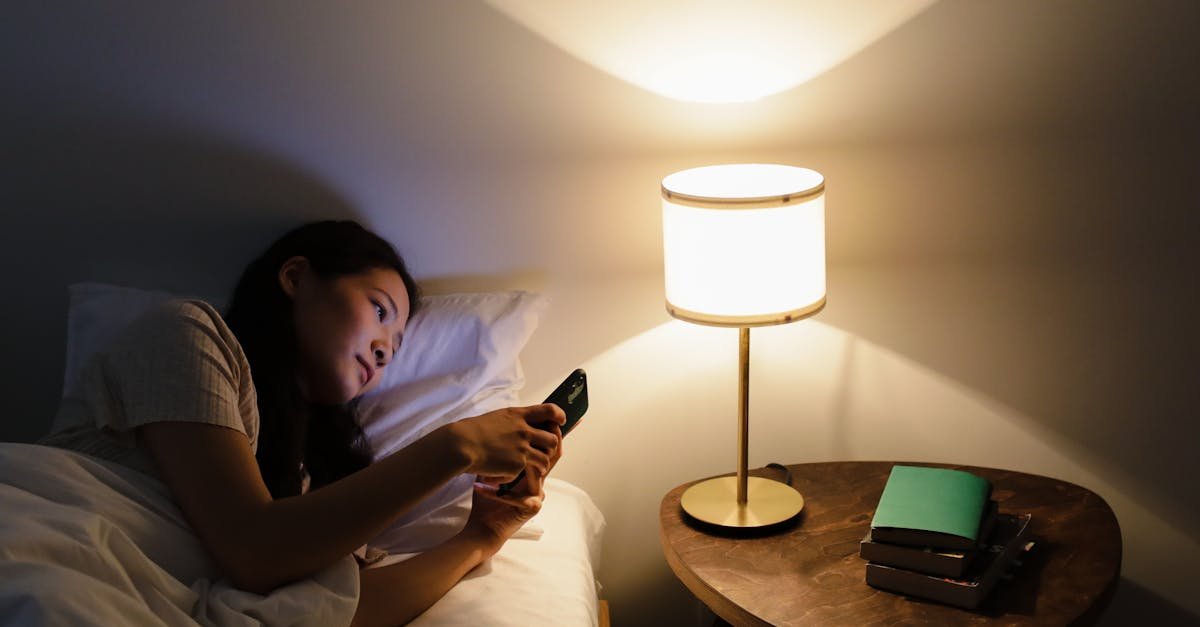 A woman relaxes in bed, using her smartphone beside a lit table lamp in a cozy room.