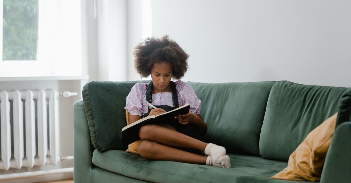 A young girl sits comfortably on a green sofa, writing in a notebook near a bright window.